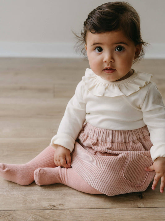 Child wearing a white top with ruffled collar and pink skirt sitting on a wooden floor. Baby Corduroy Skirt - Blush Baby girl