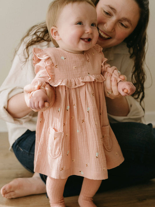 Woman holding a baby wearing a pink dress with ruffled sleeves and a pocket.