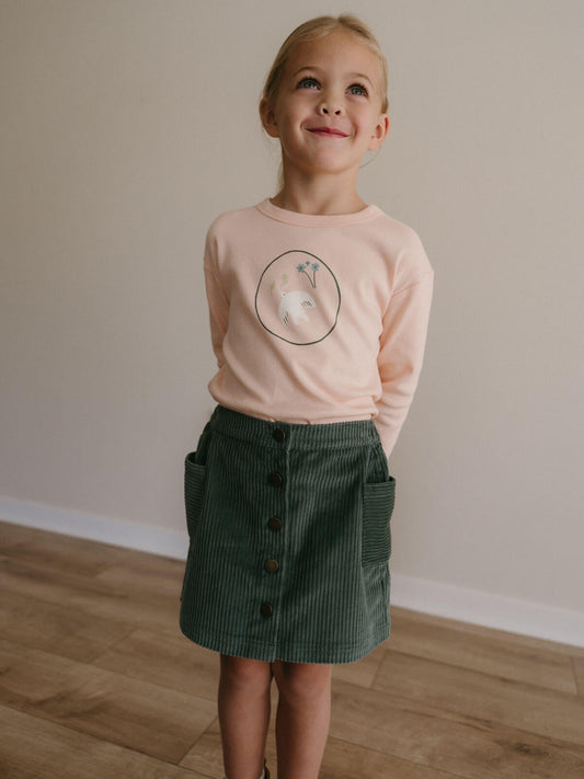 Young girl wearing a pink shirt with a graphic and a green skirt standing on a wooden floor. Corduroy Skirt - Balsam Girls