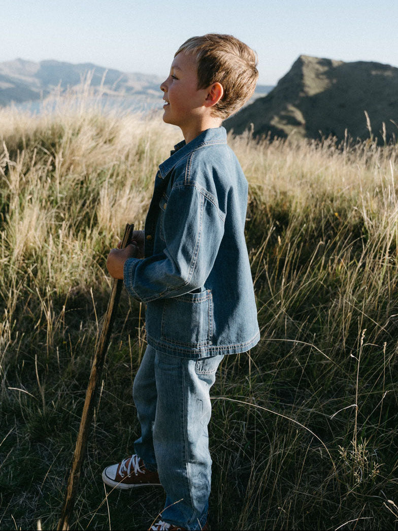 Young person in denim outfit standing in grassy field with mountains behind Denim Pants - Blue Denim Girls and Boys