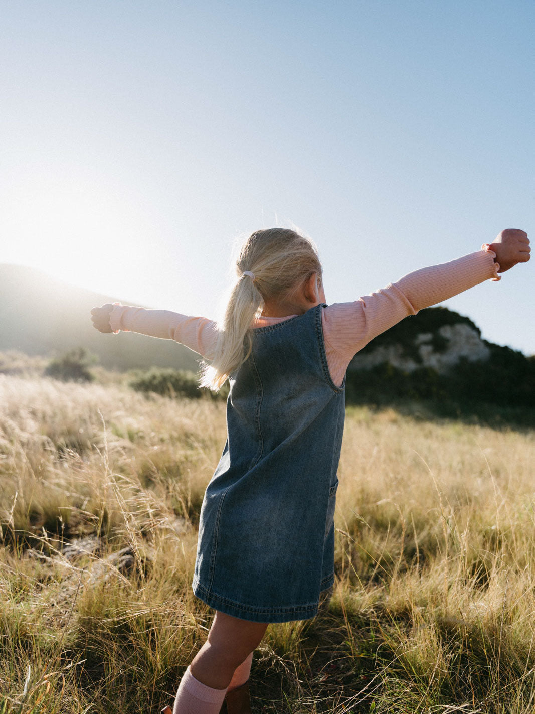 Girl in denim pinafore and pink shirt standing in grassy field with arms outstretched Denim Pinafore Dress - Blue Denim Girls