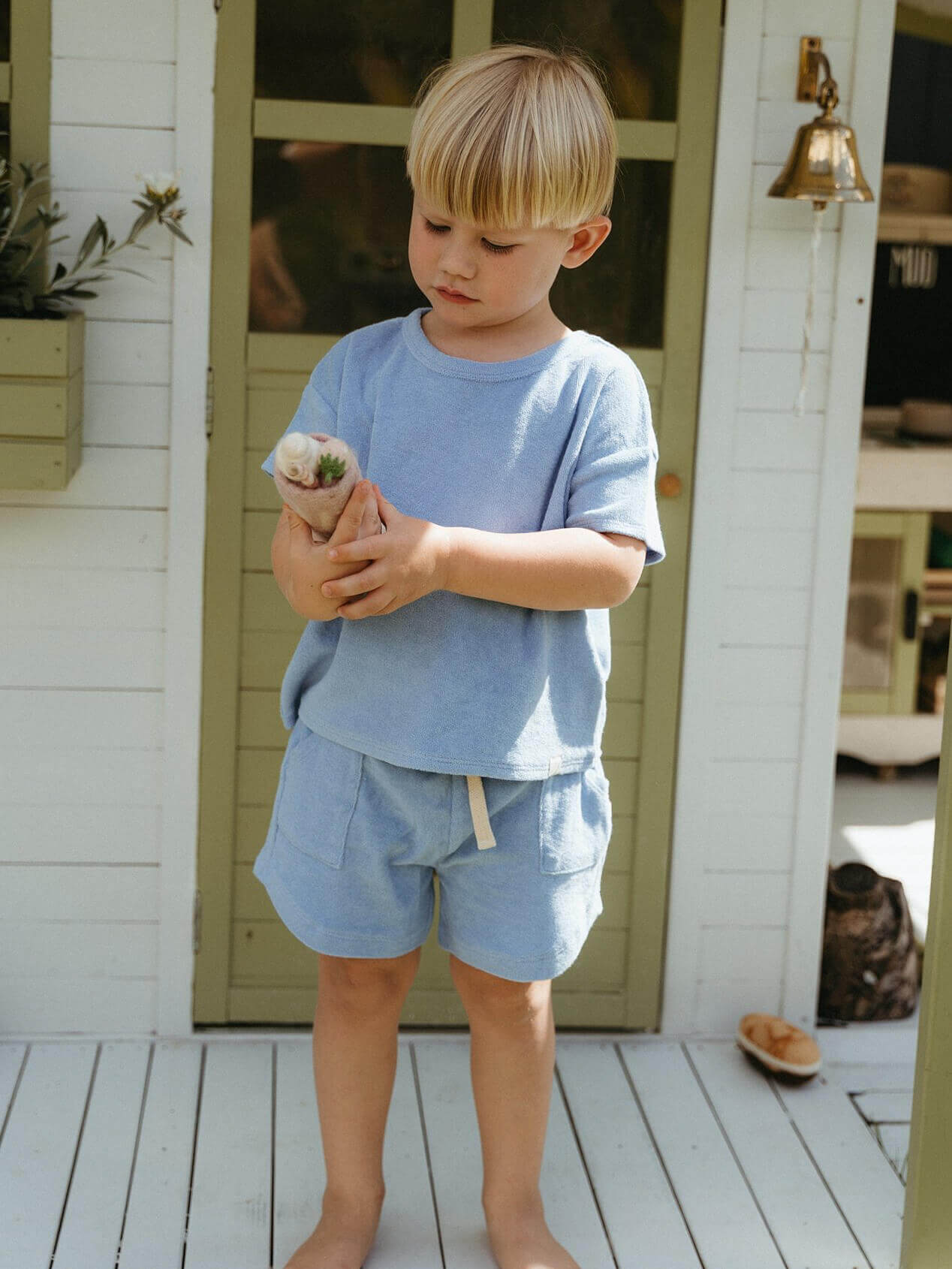 Child in light blue outfit holding a small plant on a wooden deck.