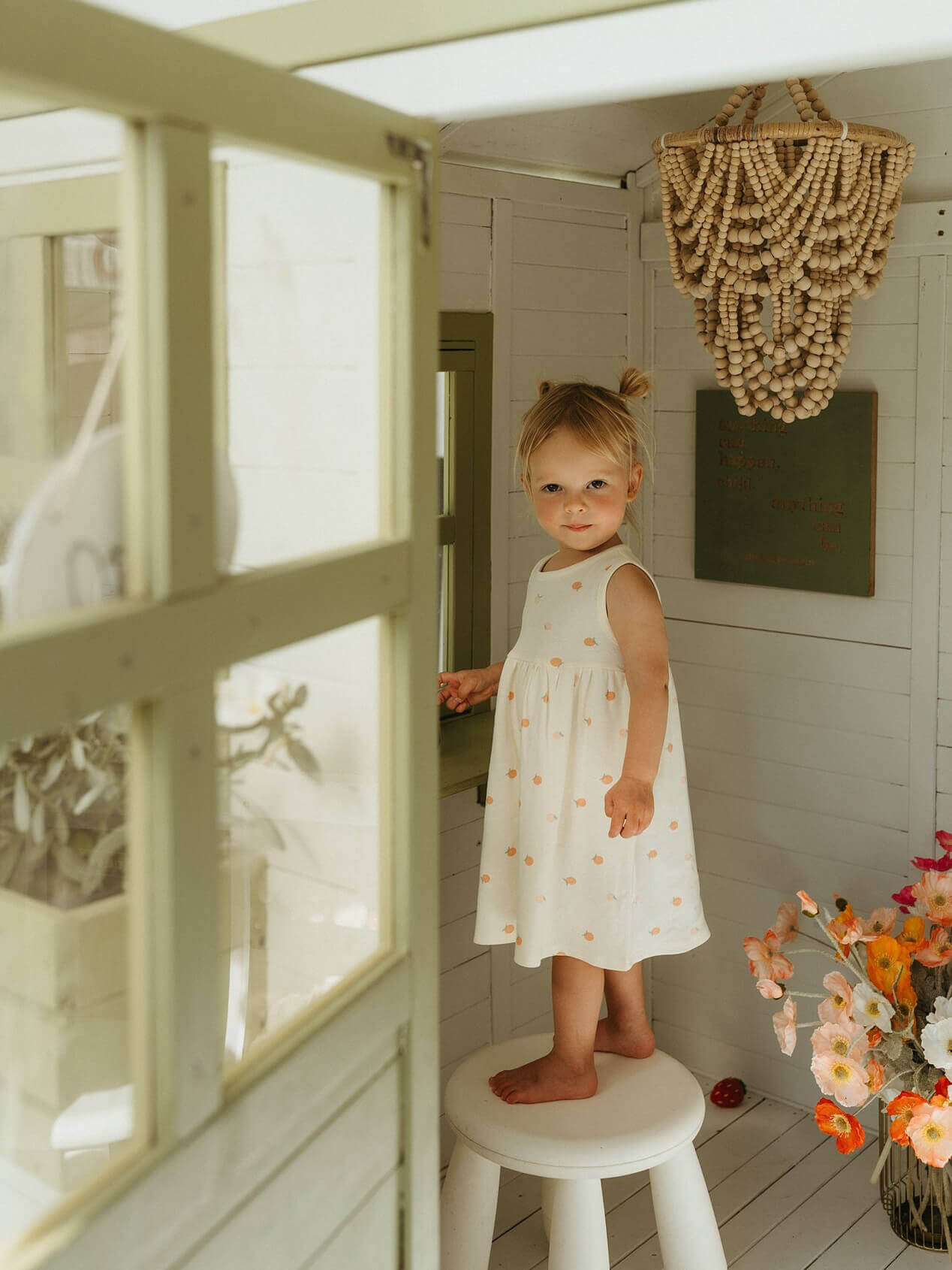 Young girl in a white dress standing on a stool in a bright room with decorative elements.