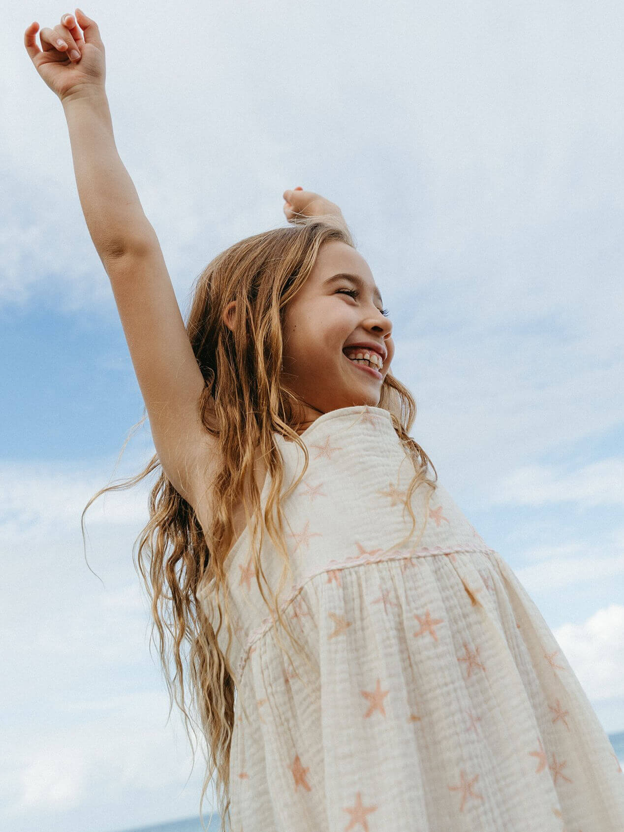 Young girl in a star-patterned dress with arms raised against a blue sky. Juliette Dress - Starfish Girls