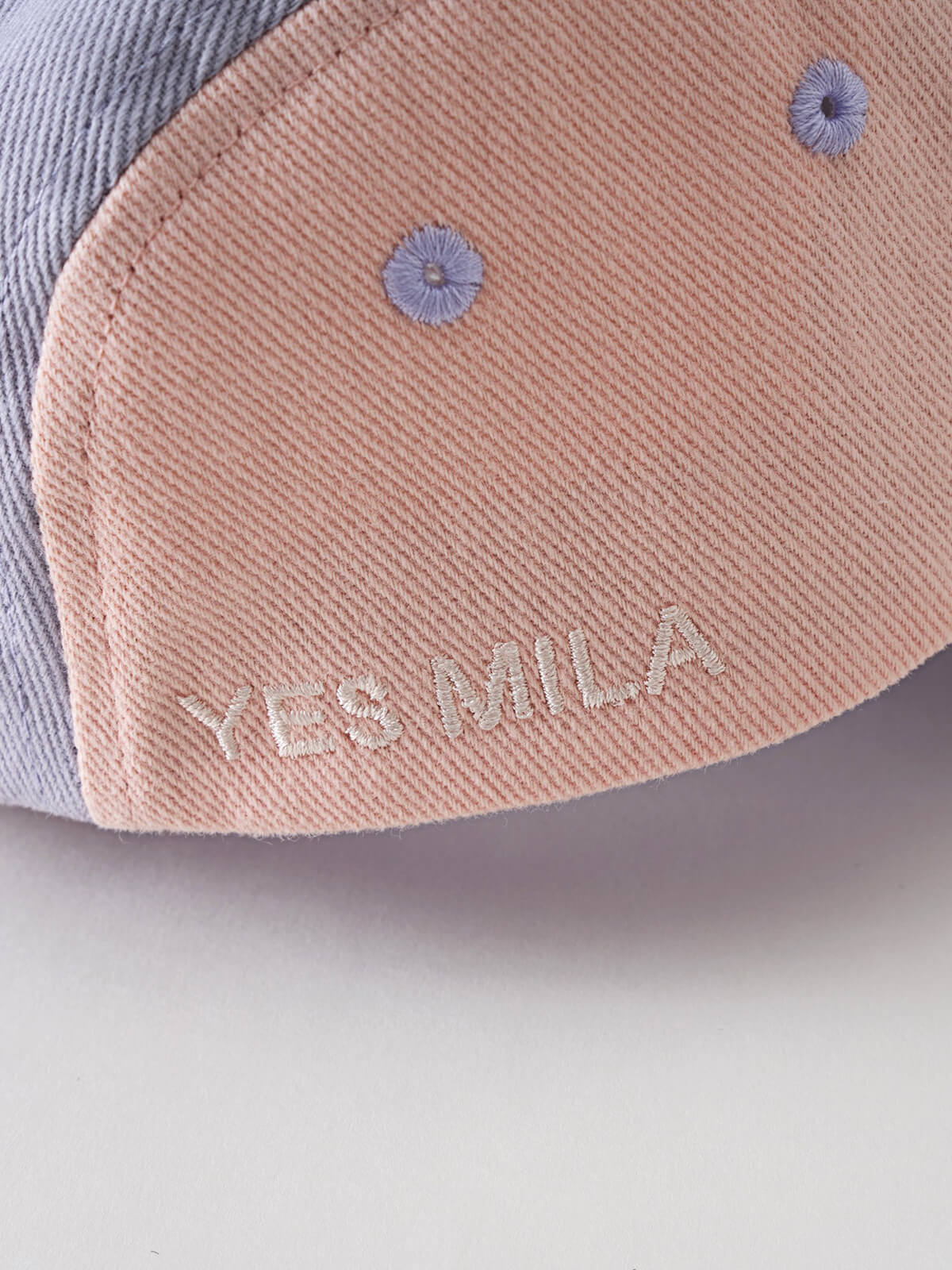 Close-up of a peach cap with blue embroidered details and text Lily Cap - Lilac Girls