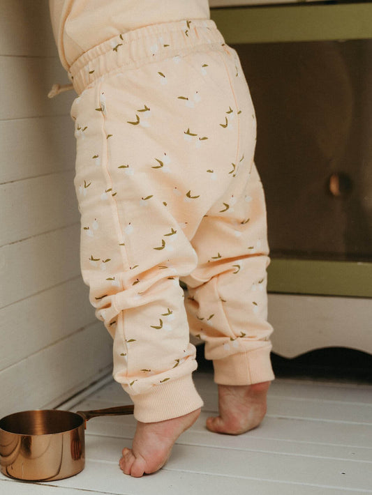 Child wearing cream-colored pants with a subtle pattern, standing on a wooden floor next to a copper pot.