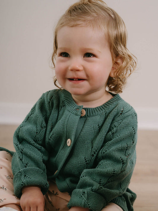 Child wearing a green knitted sweater sitting on a wooden floor. Pointelle Cardigan - Balsam Girls
