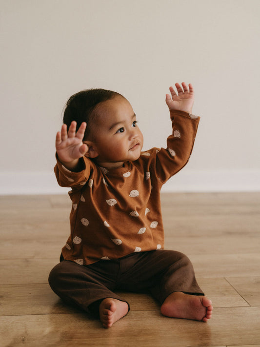 Baby sitting in a rust-coloured bear-print long-sleeved top and brown trousers.