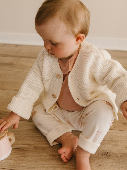 Baby wearing an ecru cotton cardigan over a pink top and patterned trousers.