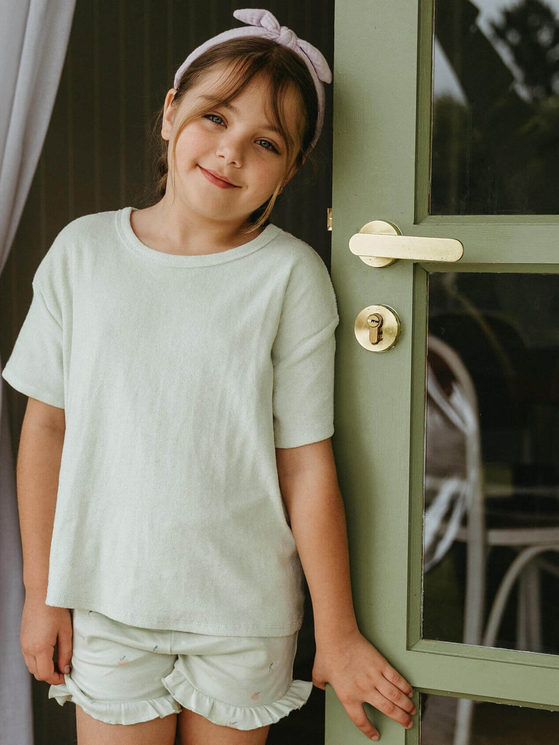 Child wearing a mint towelling tee with printed shorts beside a green door.