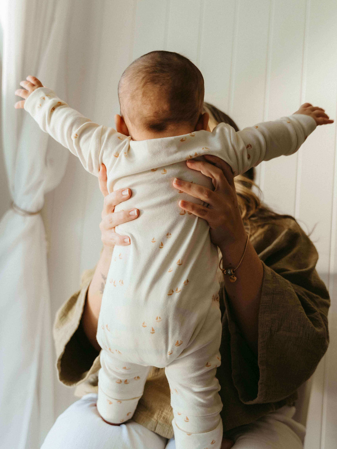Baby in a Sailing print one-piece standing on a parent’s lap with arms outstretched.