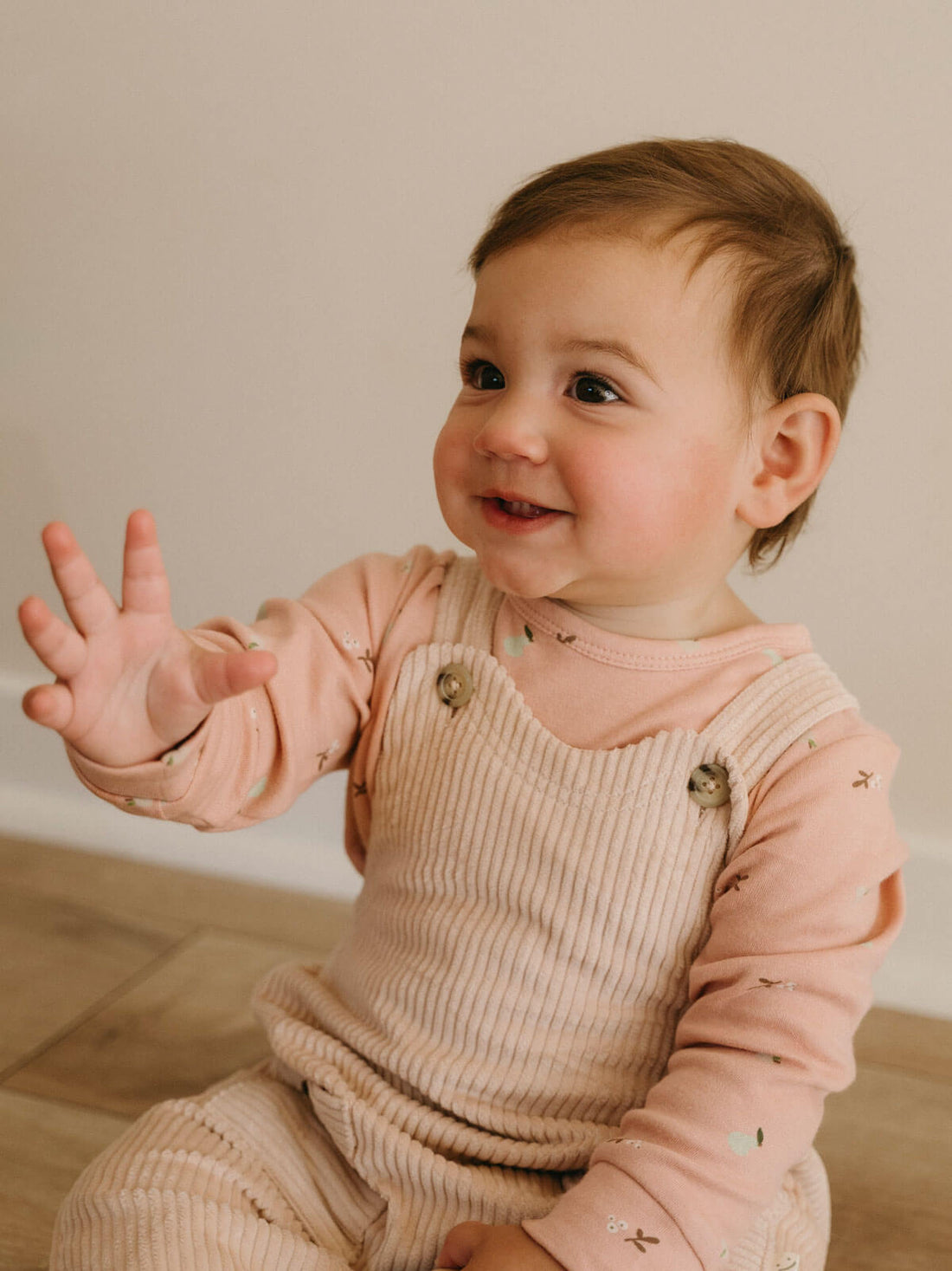 Baby sitting in Yes Mila corduroy overalls in Clay layered over a printed long sleeve tee