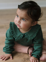 Young child in green long-sleeved top with ruffled collar sitting on wooden floor Baby Ruffle Top - Balsam Baby girl