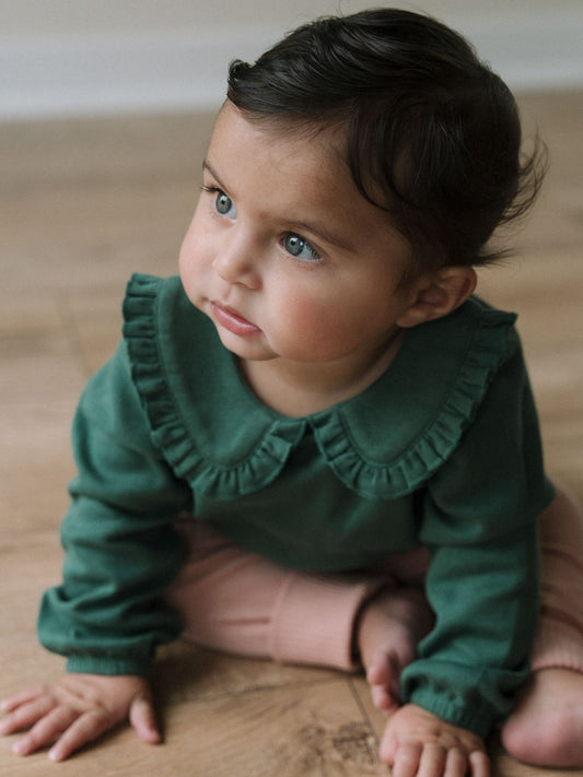 Young child in green long-sleeved top with ruffled collar sitting on wooden floor Baby Ruffle Top - Balsam Baby girl