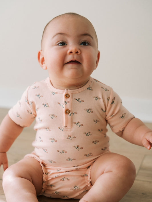 Baby sitting on white surface wearing peach floral patterned short sleeve bodysuit Henley Short Sleeve Bodysuit - Wildflower Bouquet Pink Baby girl