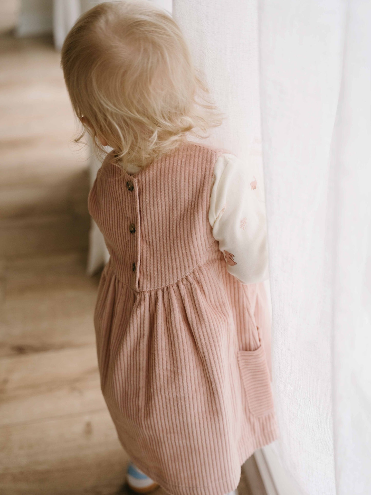 Child wearing a pink corduroy dress standing indoors with a neutral background