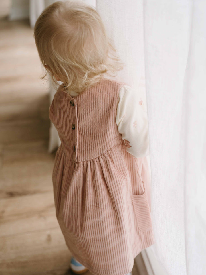 Child wearing a pink corduroy dress standing indoors with a neutral background