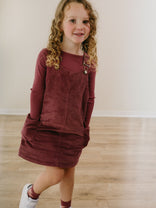 Young girl wearing burgundy corduroy pinafore dress and long-sleeved shirt Corduroy Pinafore Dress - Plum Jam Girls