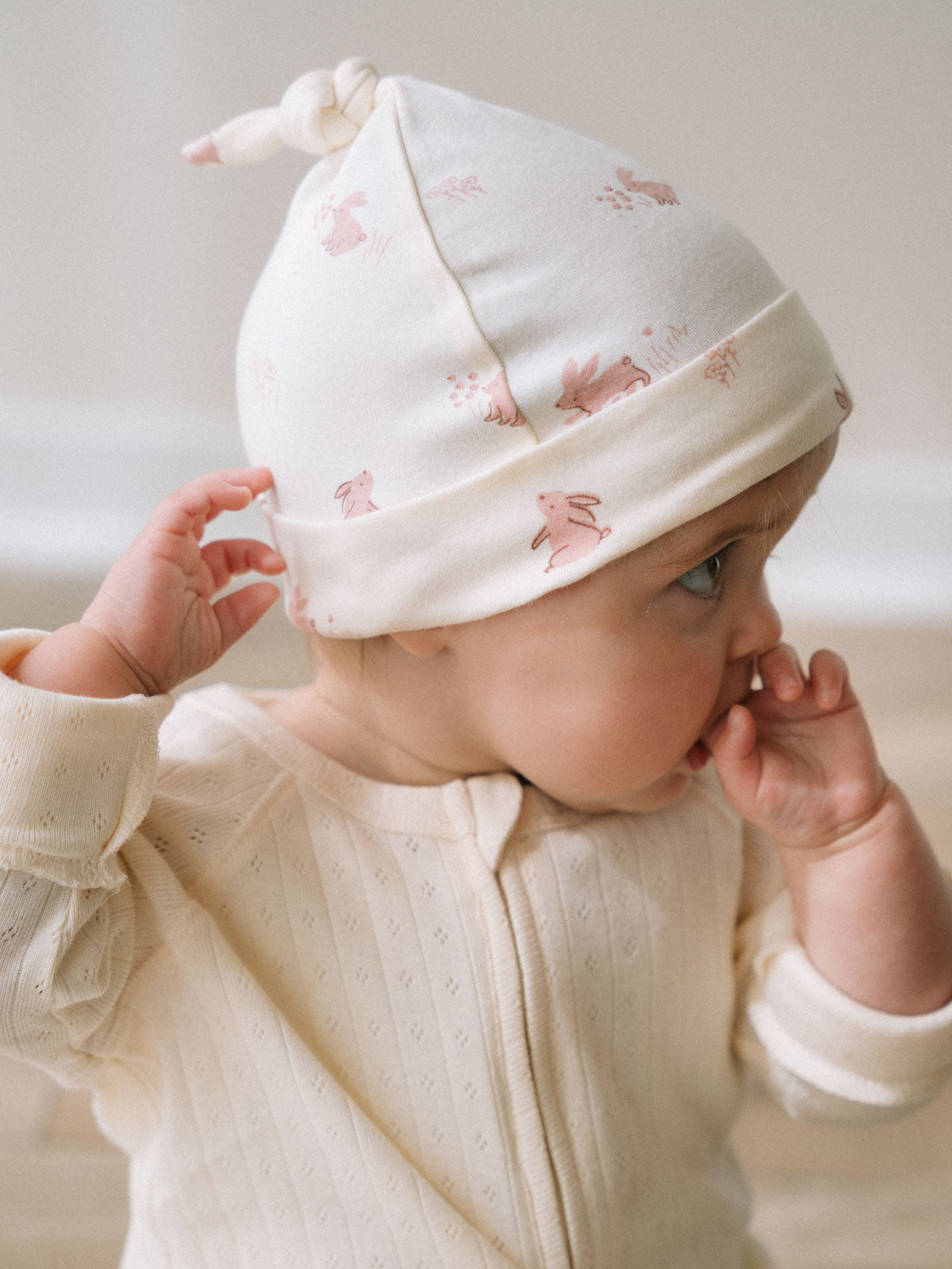 Baby wearing a white hat with pink bunny print, hand touching brim Knot Hat - Meadow Friends Ecru Baby girl