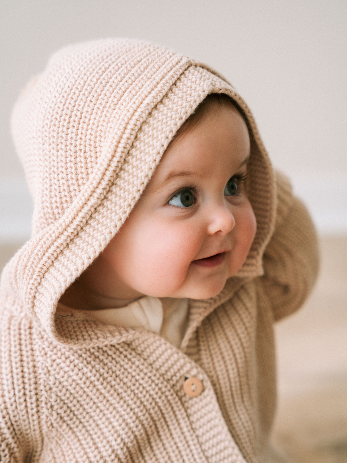 Baby wearing a beige knitted hoodie with a hood, looking to the side.