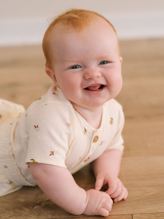 Baby with red hair smiling on wooden floor wearing patterned short sleeve bodysuit Henley Short Sleeve Bodysuit - Autumn Harvest Ecru Baby girl