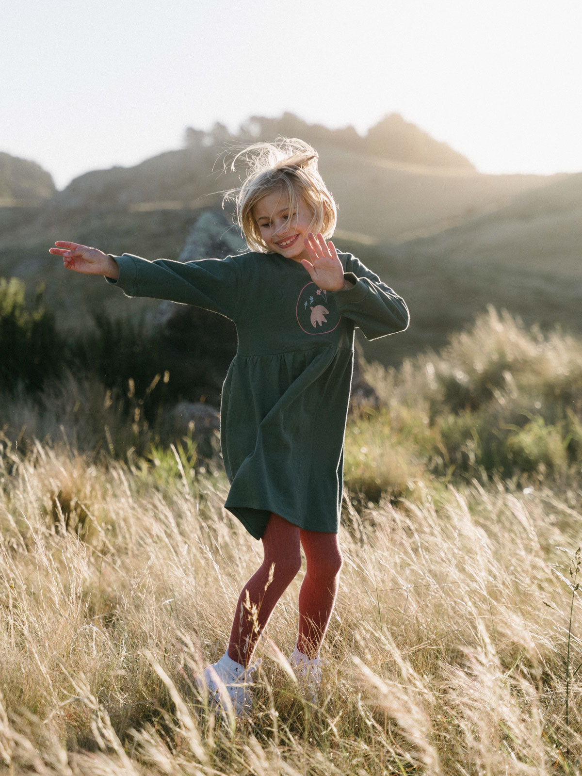 Young girl jumping in a grassy field wearing a green sweatshirt dress Sweatshirt Dress - Dove Girls