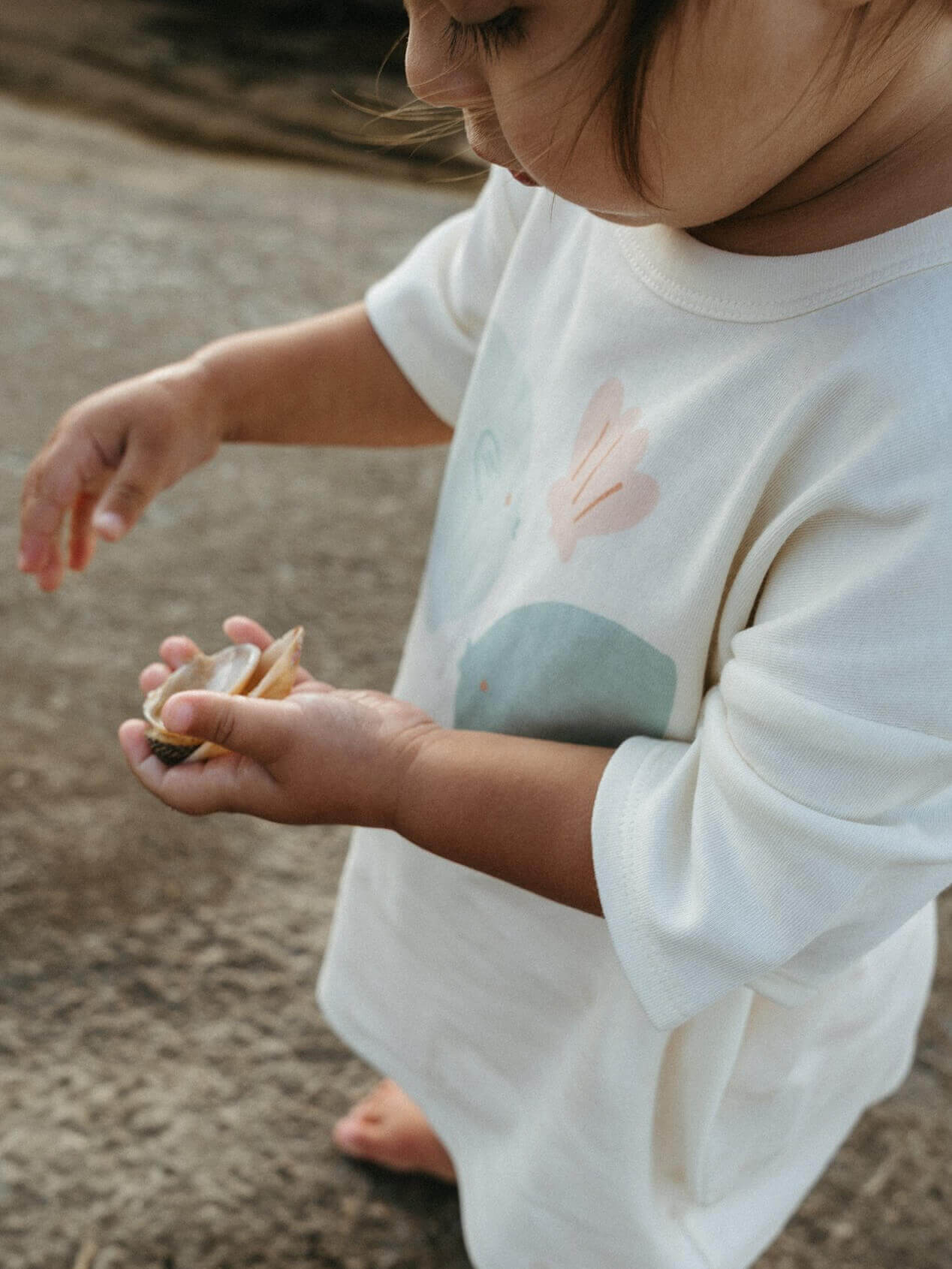 Child holding a shell on a sandy beach wearing a white shirt with nature-themed design.
