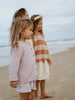 Two young girls standing on a beach, wearing light clothing.