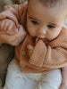 Baby wearing a knitted brown sweater holding onto a person's hand.