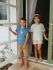 Two children standing on a porch with a white door and window in the background.