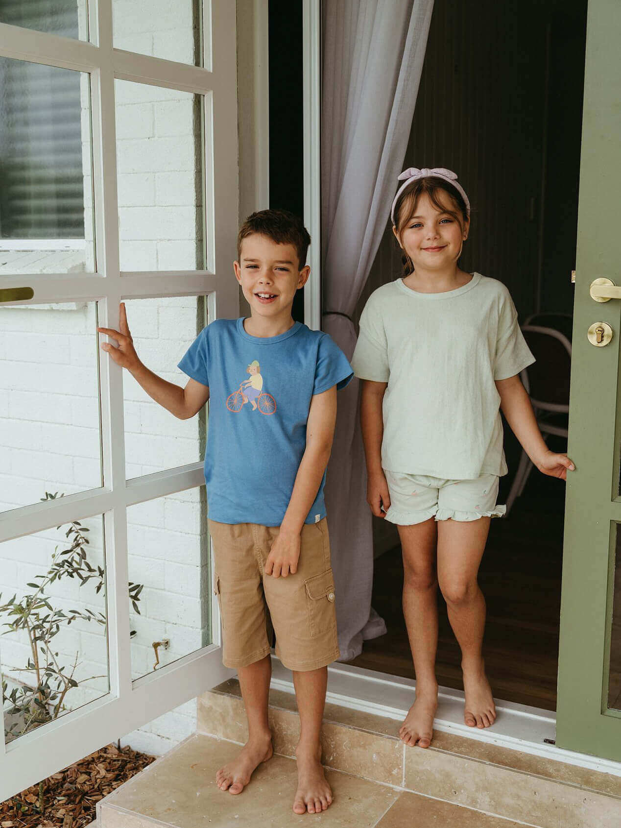 Two children standing on a porch with a white door and window in the background.
