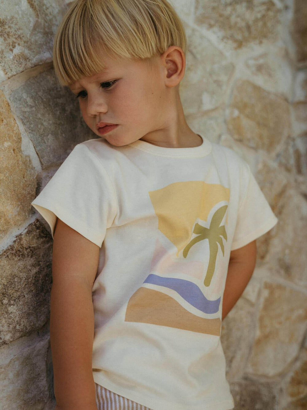 Child wearing a t-shirt with a palm tree design against a stone wall.