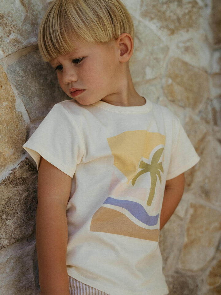 Child wearing a t-shirt with a palm tree design against a stone wall.