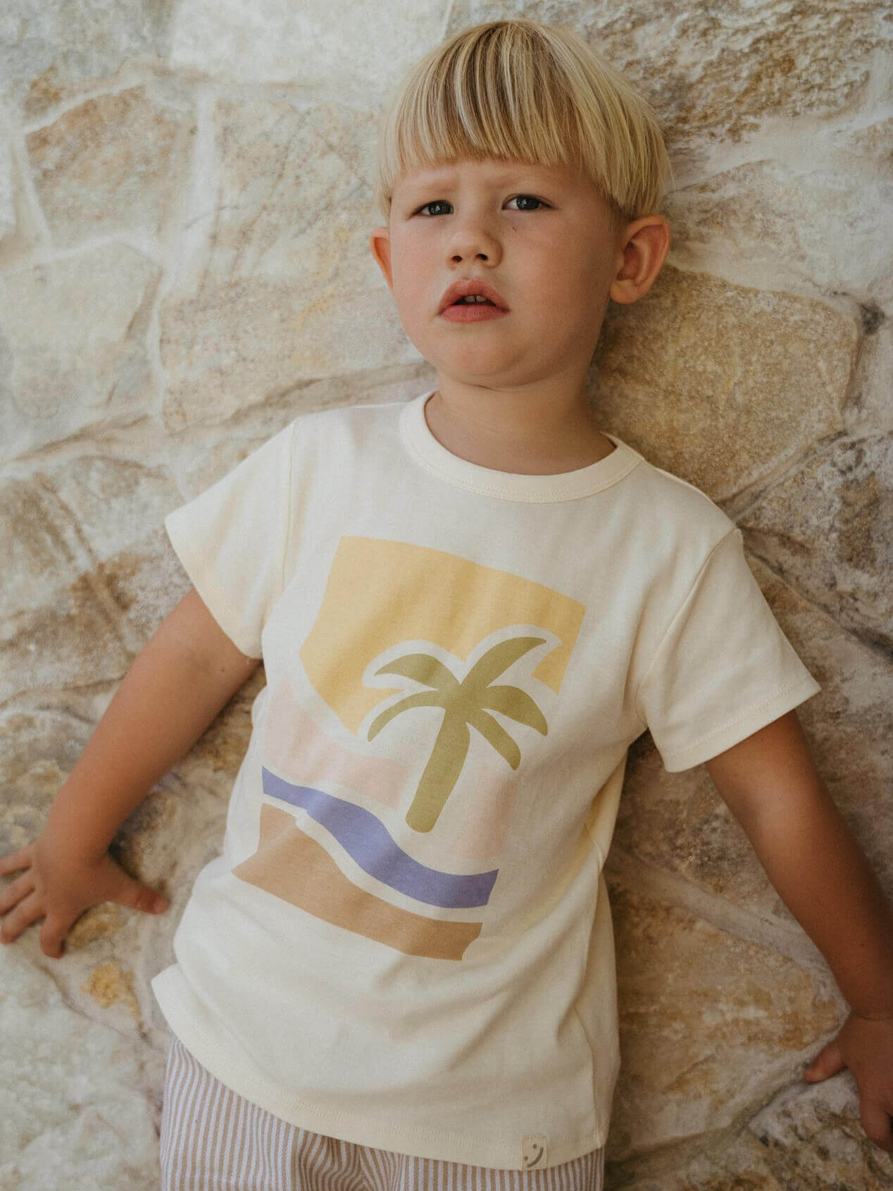 Child wearing a t-shirt with a palm tree design against a stone wall.