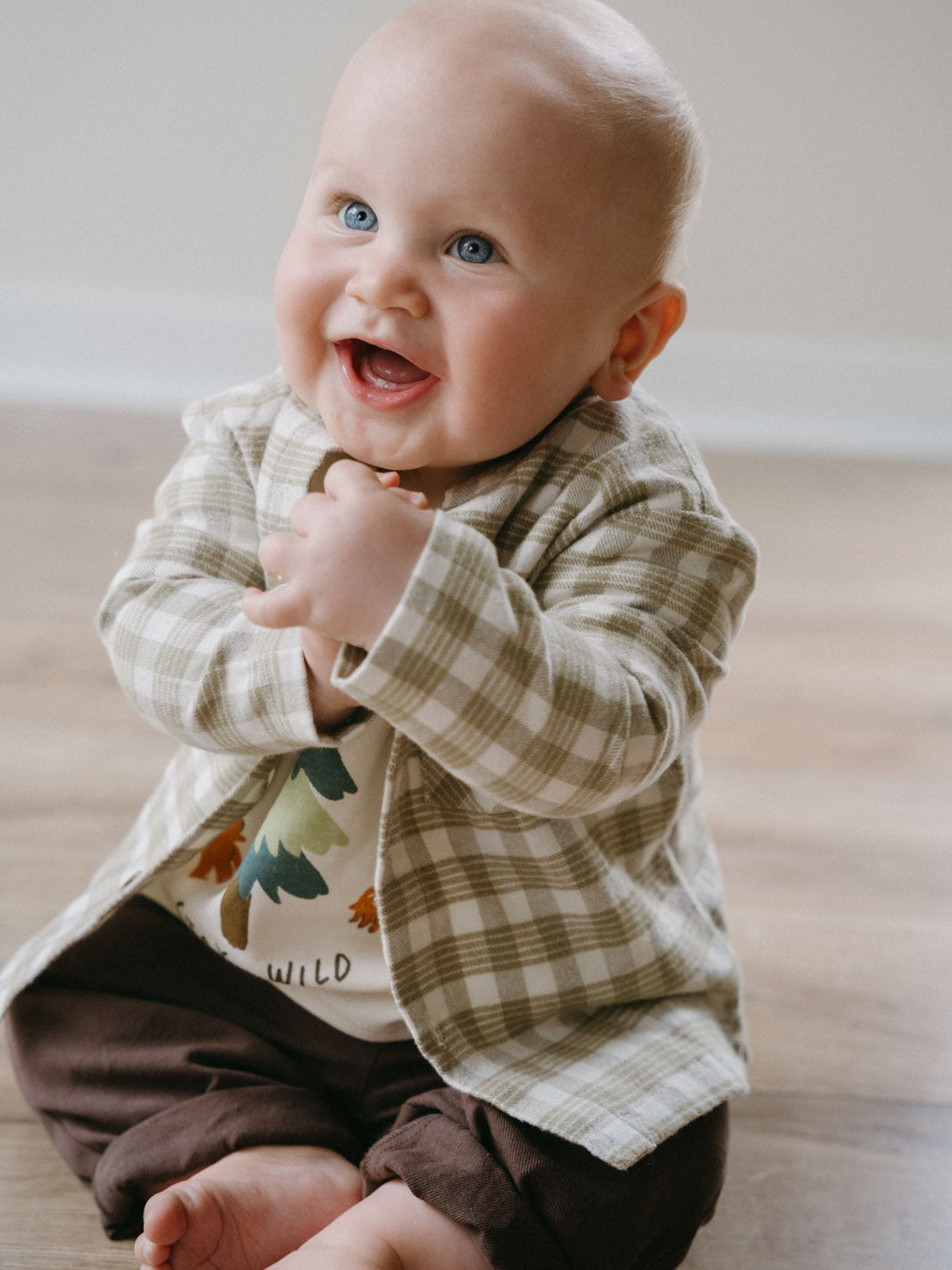 Baby wearing plaid shirt and brown pants sitting on floor smiling Baby Check Shirt - Woven Check Baby boy