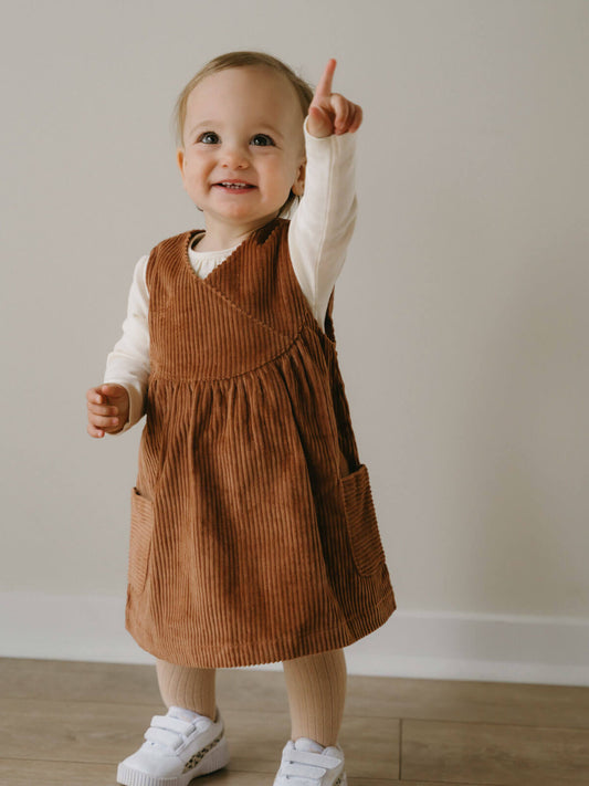Child wearing a brown corduroy dress with a white shirt underneath, standing on a wooden floor.