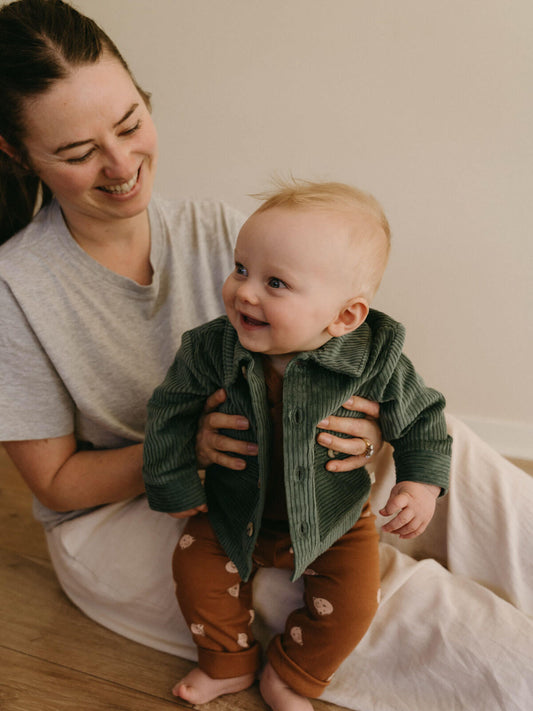 Woman holding a baby wearing a green jacket and brown pants in a home setting.