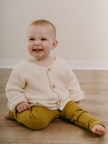 Baby wearing a cream cardigan and mustard pants sitting on a wooden floor.