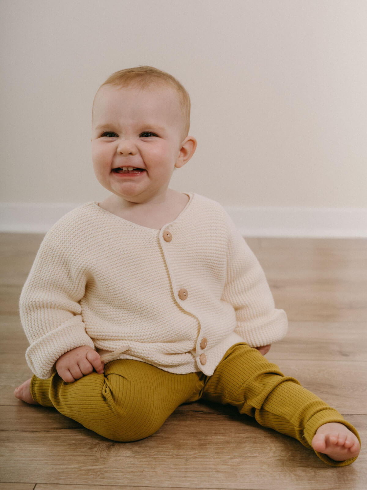 Baby wearing a cream cardigan and mustard pants sitting on a wooden floor.