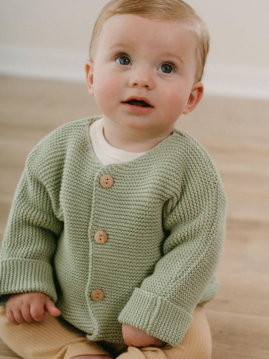 Baby wearing a green knitted cardigan with brown buttons on a wooden floor.