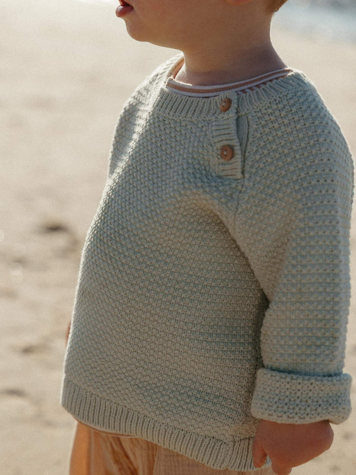 A close-up side view of a child wearing a textured knit sweater on a sandy beach. The sweater is a light oatmeal-beige color with a small button detail on the chest.