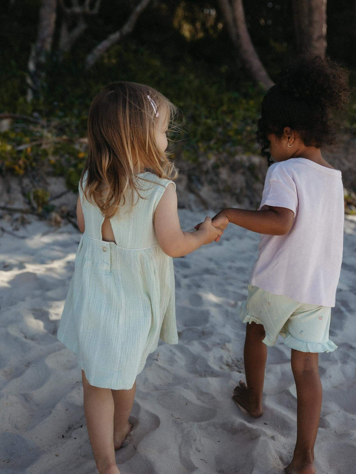 Two children holding hands walking on a sandy beach, viewed from behind. The child on the left wears a pale mint green sleeveless dress, and the child on the right wears a light pink t-shirt and pale green shorts.