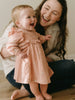 Woman laughing with a baby in a pink dress on a wooden floor. Baby Woven Dress - Autumn Harvest Rosehip Baby girl