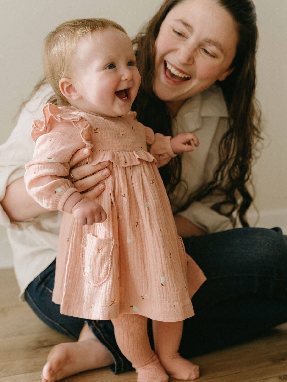 Woman laughing with a baby in a pink dress on a wooden floor. Baby Woven Dress - Autumn Harvest Rosehip Baby girl