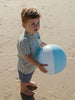A young boy standing on a sandy beach holding a light blue and white beach ball. He is wearing a light blue and white vertically striped short-sleeve shirt and blue shorts.