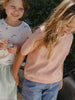 Two children standing outdoors, one in a light pink sweater and the other in a white shirt with patterns.