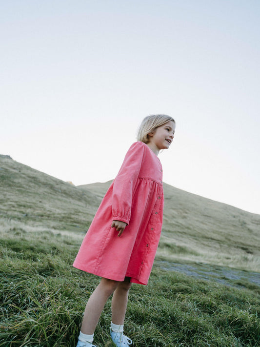 Girl wearing a pink dress standing on a grassy hill with a clear sky.