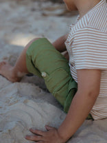 A child sitting on sandy ground wearing olive green cargo shorts and a cream and brown striped t-shirt.