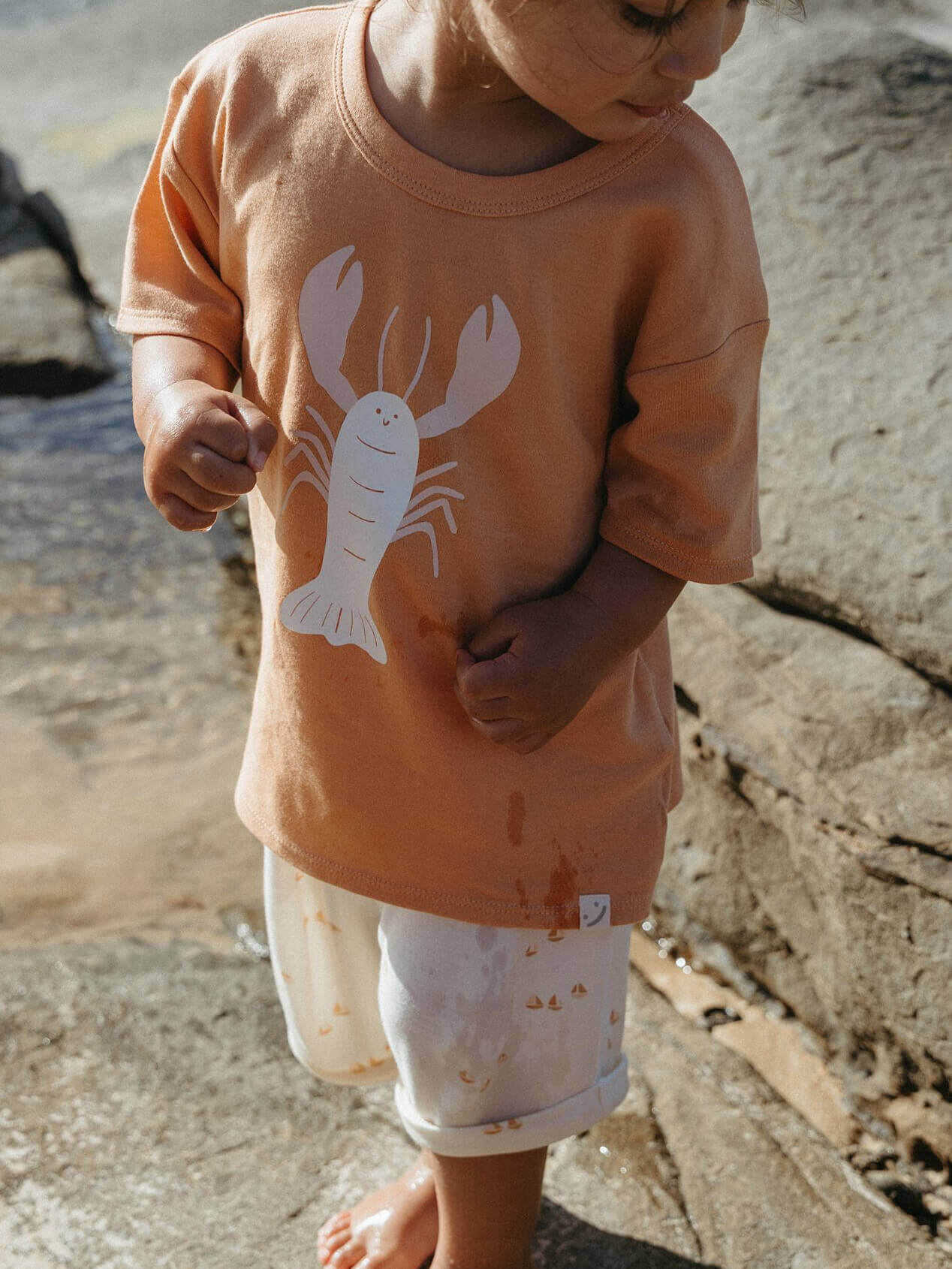 Child wearing a peach-colored shirt with a lobster design outdoors on a rocky surface.