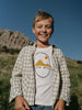 Child wearing a 'Stay Wild' t-shirt in a natural setting with mountains.